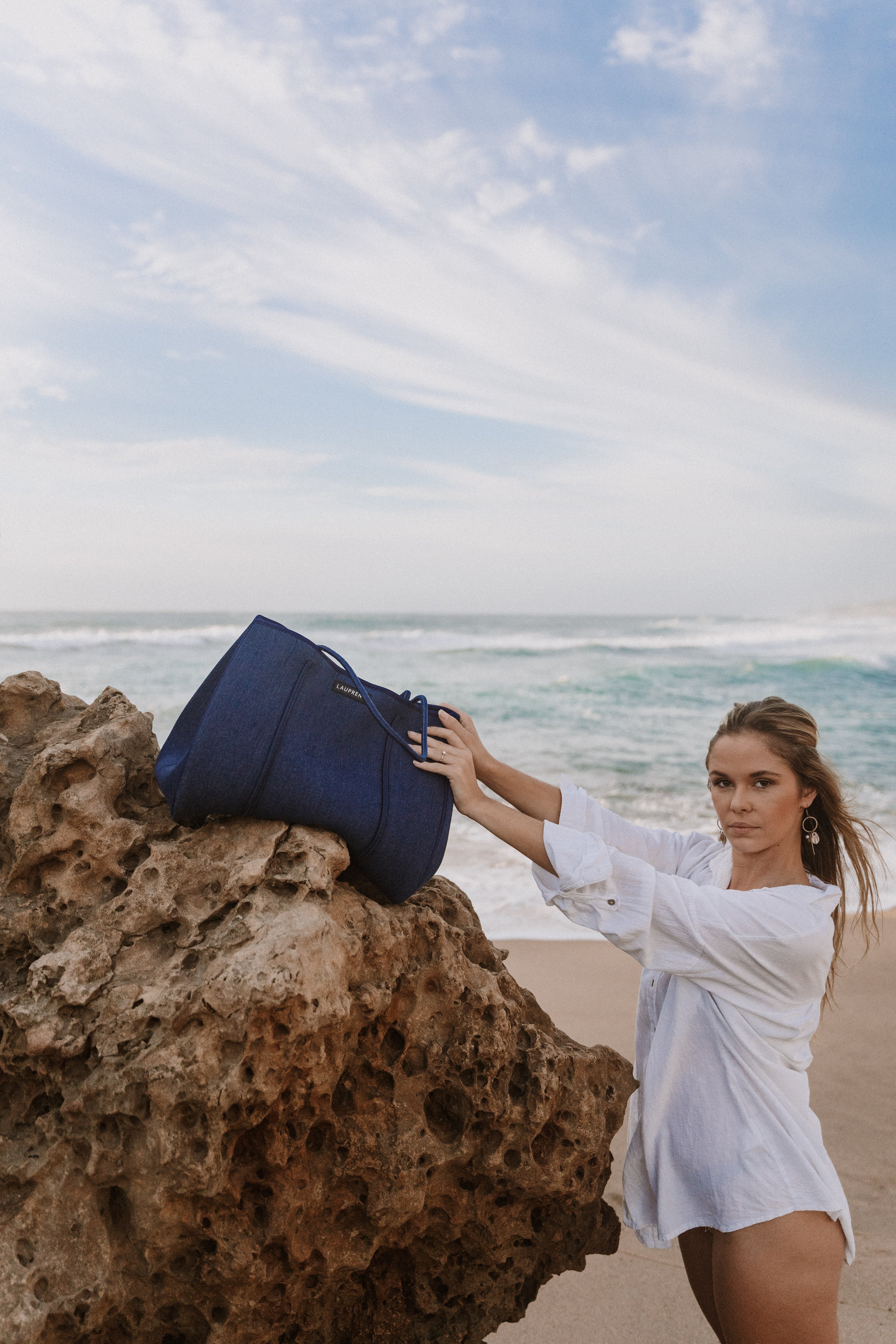 Woman in a white dress holding a blue bag on a rocky beach with ocean and sky in the background